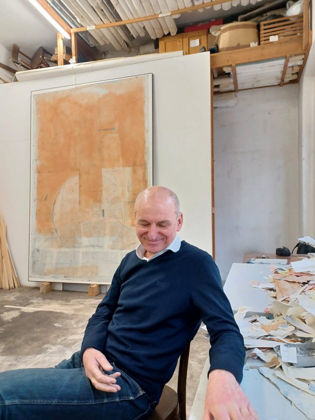 Christoph Luger (schlank, blauer Pulli, zartes Gesicht, wenig Haare) sitzt auf einem Sessel in seinem Atelier vor einer Wand mit einem unfertigen Bild.